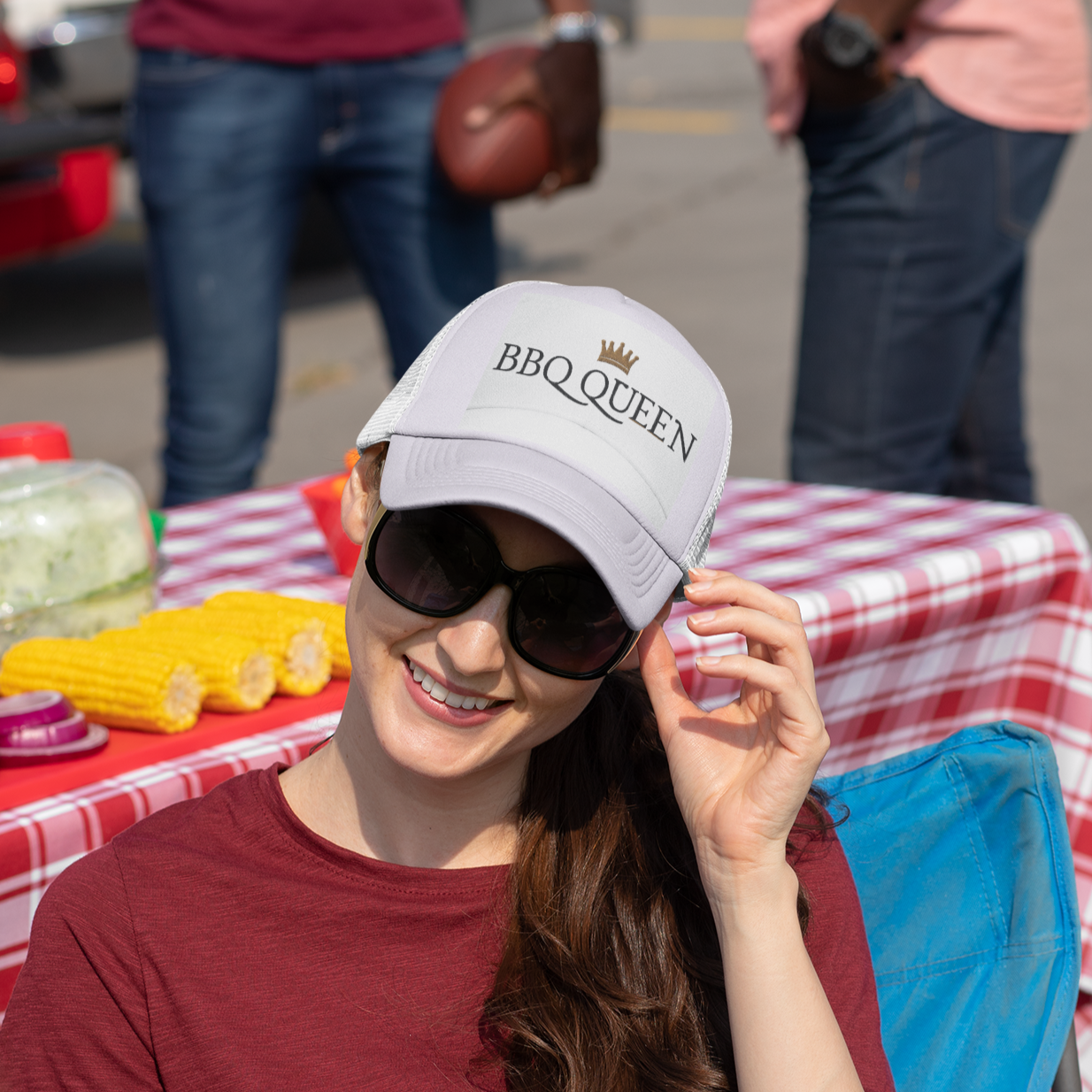Woman wearing a 'BBQ Queen' cap sitting at a table with food in an outdoor setting alpha bbqear cap